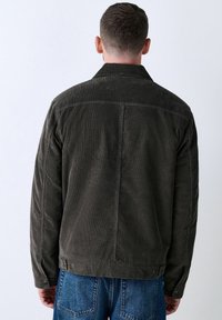 Hombre vestido con una chaqueta de pana gris oscuro y jeans azules, visto de espaldas contra un fondo claro y liso.