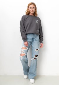 Grauer Sweatshirt mit einem Logo auf der Brust, kombiniert mit hellblauen, verwaschenen Jeans mit großen Rissen und einem lockeren Sitz. Weiße Sneakers runden das Outfit ab.