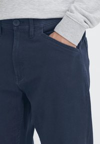 Temno modre bombažne hlače s teksturirano tkanino, tradicionalnim dizajnom s petimi žepi in zapiranjem v pasu na gumbe.