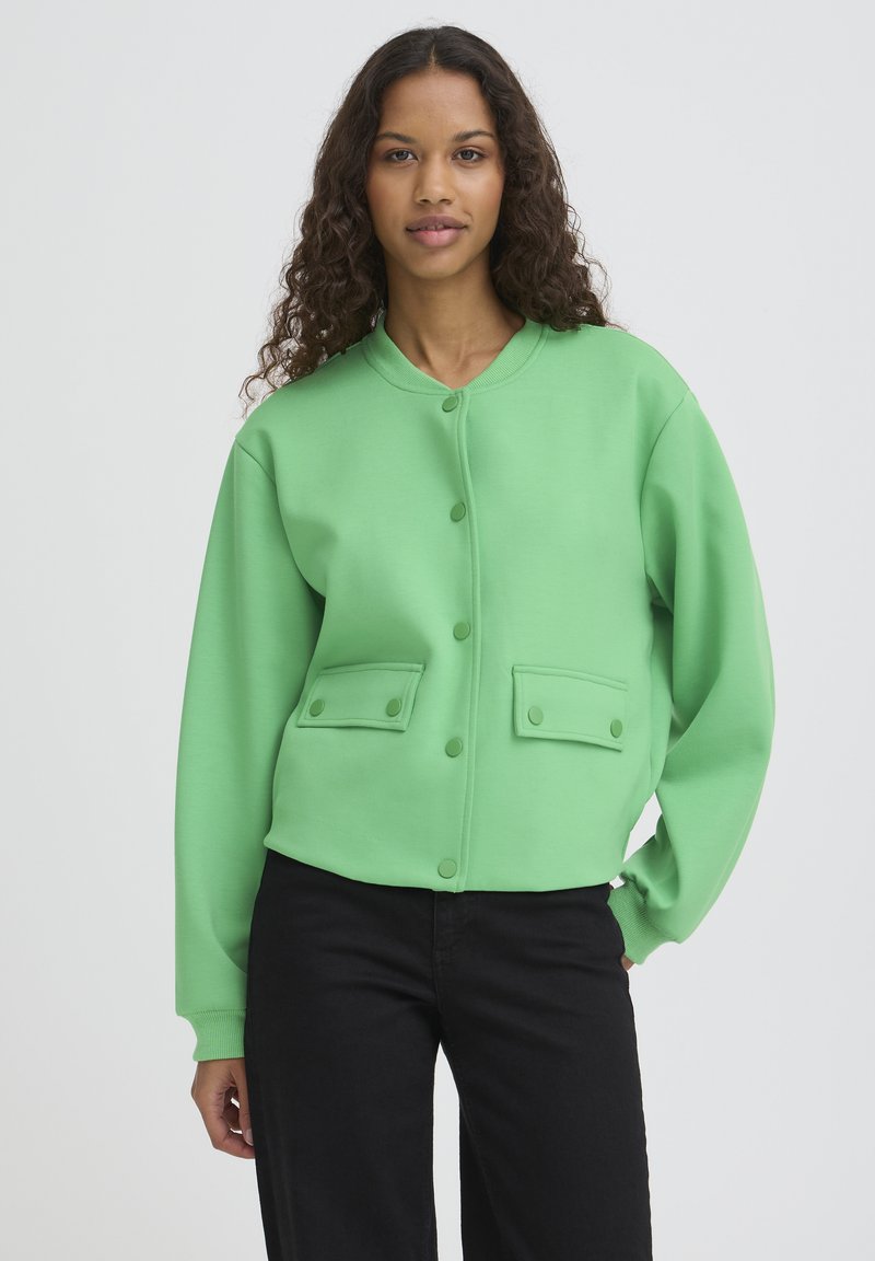 Zelena majica z okrolim izrezom, zapenjanjem na gumbe in dvema žepoma spredaj. Ima dolge rokave in gladek, teksturiran material.