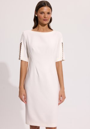 Kobieta w białej sukience do kolan z rękawami do łokcia, ozdobionymi perełkowymi guzikami wzdłuż rozcięć na rękawach.