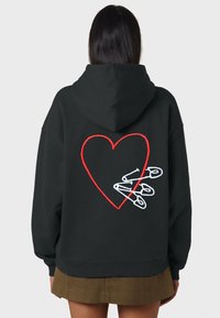 Čierna mikina s kapucňou, na ktorej chrbte je veľké červené vyšívané srdce obklopené bielymi bezpečnostnými špendlíkmi. Materiál vyzerá mäkko a má voľný strih.