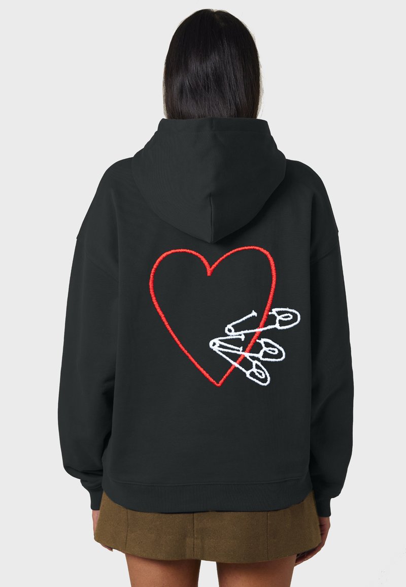 Čierna mikina s kapucňou, na ktorej chrbte je veľké červené vyšívané srdce obklopené bielymi bezpečnostnými špendlíkmi. Materiál vyzerá mäkko a má voľný strih.