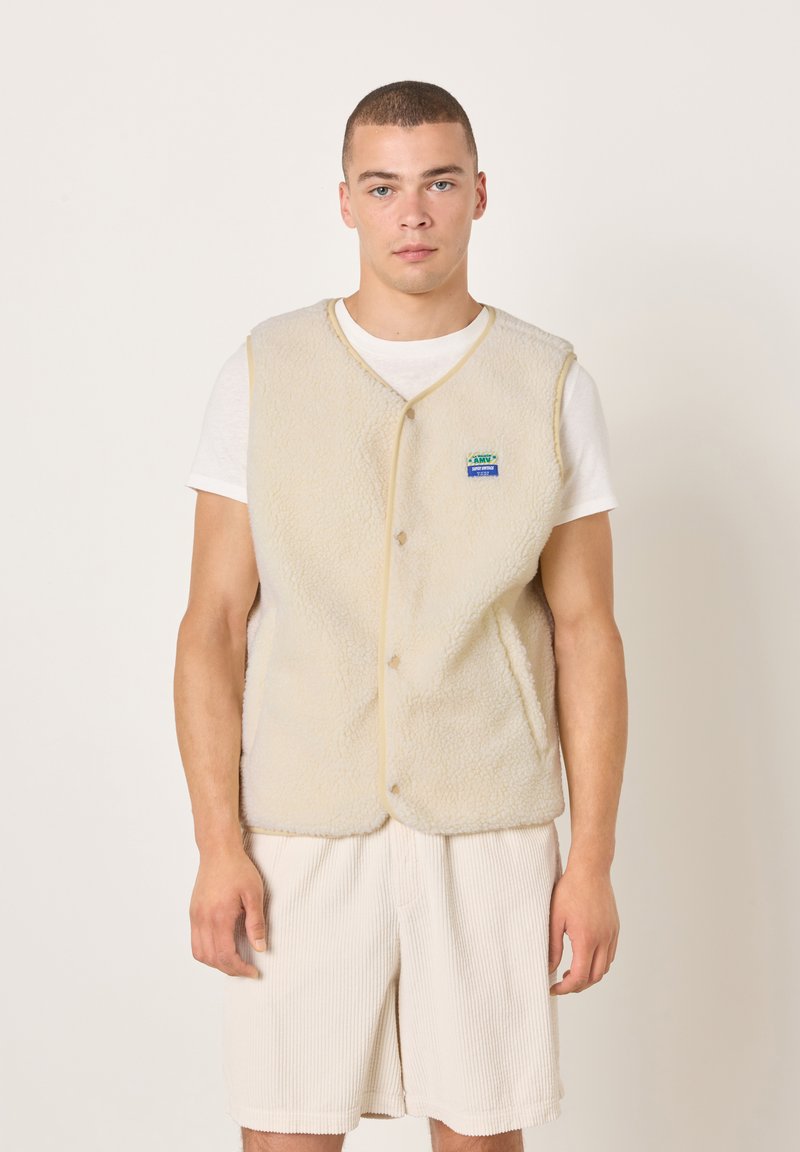 Smėlinio spalvos vilnonė liemenė su sagomis priekyje, apvalia iškirpte ir mažu mėlynu bei žaliu logotipu ant kairiojo krūtinės šono. Dėvima ant balto marškinėlio.