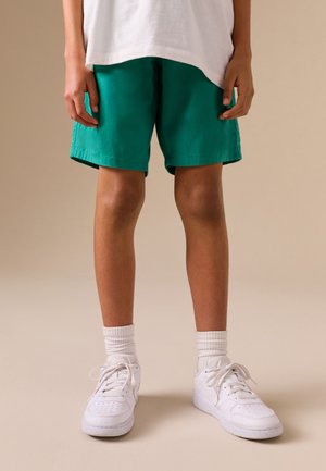 Kind trägt grüne Shorts, weiße Sneakers und weiße Socken und steht vor einem schlichten beigen Hintergrund.