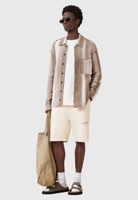 Camicia a righe color tan e beige con taschino, t-shirt bianca, pantaloni corti beige, sandali e una borsa a tracolla marrone. Gli accessori includono occhiali da sole scuri.