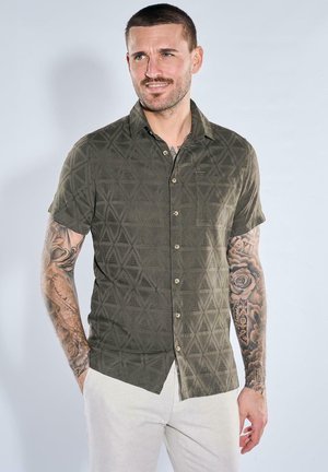 Mann mit kurzen Haaren und Tätowierungen auf beiden Armen, der ein grünes, gemustertes Kurzarmhemd und helle Hosen trägt, leicht lächelnd.