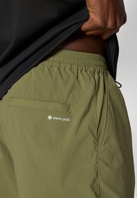 Hand lifting black shirt to reveal olive green elastic-waist shorts with back pocket and white "snow peak" logo.