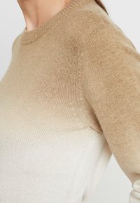 Beige graduerande tröja med rund halsringning, ribbade ärmslut och mjukt, texturerat stickat material. Färgen går från mörk till ljus.