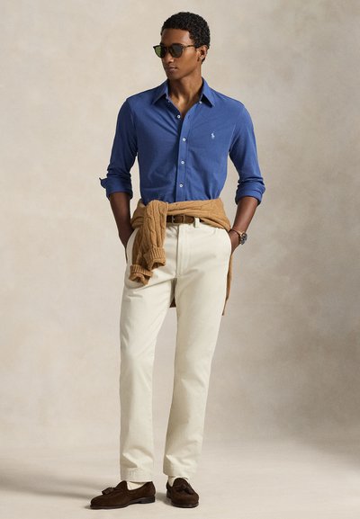 Μπλε πουκάμισο με κουμπιά, κρεμ chino παντελόνι, καφέ πλεκτό πουλόβερ δεμένο στη μέση, σκούρα παπούτσια και γυαλιά ηλίου. Λεπτό ύφασμα και άνετη εφαρμογή.