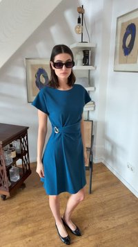 Femme portant une robe bleu sarcelle avec une boucle latérale et des ballerines noires, debout à l'intérieur près d'une œuvre d'art abstraite encadrée et d'une étagère.