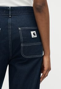 Tmavomodré rifľové nohavice s bielym prešívaním a zadným vreckom so štítkom značky Carhartt, nosené osobou s tmavou pokožkou.