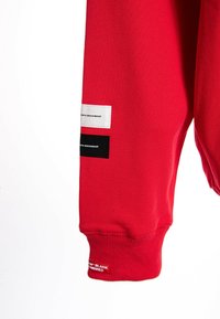 Sudadera roja con puños acanalados y tres etiquetas en la manga en blanco y negro, que presentan el texto "movimiento de ola". Textura de tela suave.