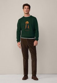 Sudadera verde oscura con un diseño de perro ilustrado en naranja y azul, combinada con pantalones de pana marrones y zapatos marrones con cordones.
