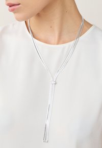 Silberne Halskette mit mehreren Strängen und einem zentralen Knoten, die sich elegant um den Hals legt, vor einem weißen Outfit-Hintergrund präsentiert.