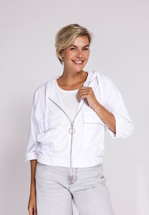 Smiling woman with short blonde hair wearing a white zip-up hoodie over a white top and light gray jeans, standing against a plain background.