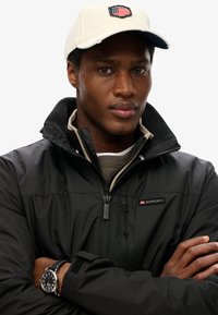 Czarna kurtka puchowa z pionowymi paskami, zapinana na zamek, z markowymi akcentami. Beżowa czapka z haftowaną flagą USA. Zegarek ze stali nierdzewnej.