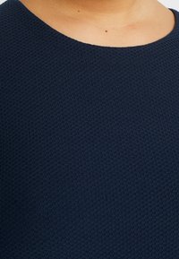 Pull-over bleu marine en tissu texturé à motif alvéolé. Encolure ronde ; coutures lisses et régulières ; légère brillance visible à la surface.