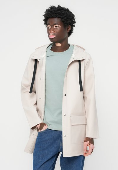 Beige vanntett jakke med hette, svarte snorer, knappelukking og lommer foran. Brukt over en mintgrønn t-skjorte og blå jeans.