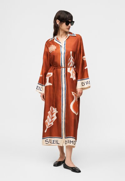 Γυναίκα που φοράει μακρύ φόρεμα σε χρώμα σκουριάς με λευκά και μπλε σχέδια, με ζώνη στη μέση, μαύρες φλατ παπούτσια και μεγάλα μαύρα γυαλιά ηλίου, όρθια.