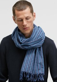 Blue plaid scarf made of lightweight fabric with fringed edges, draped around the neck, featuring varying shades of blue.