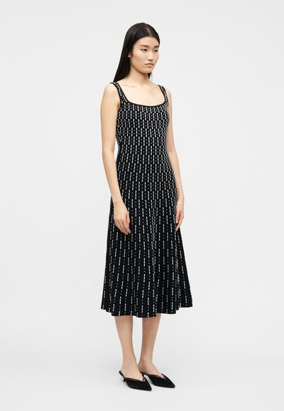 Femme portant une robe midi sans manches noire avec des lignes ponctuées blanches verticales, assortie à des ballerines slingback noires à bouts pointus, debout devant un fond blanc.