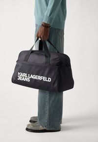 Black nylon duffle bag with dual handles, large white "KARL LAGERFELD JEANS" logo, zip closure, and structured rectangular shape.