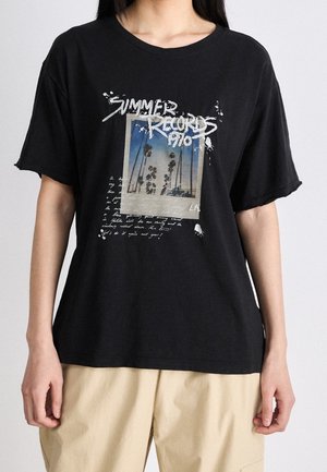 Black oversized t-shirt with a graphic of palm trees under blue sky, text "SUMMER RECORDS 1970" and handwritten-style white text, worn by person.