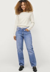 Světle béžový top s dlouhým rukávem a modré džíny s vysokým pasem a rovnými nohavicemi. Tkanina vypadá měkce a celý outfit je neformální a jednoduchý.