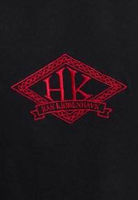 Musta kangas, jossa on punainen kirjailtu logo kirjaimilla "HK" ja teksti "HAN KØBENHAVN", kehystetty timantinmuotoisella yksityiskohtaisella koristeella.