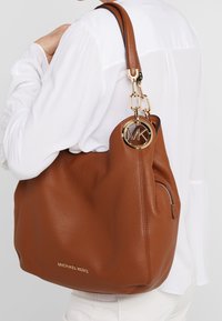 Sac hobo en cuir marron avec une surface texturée, un charm logo MK en ton doré et une poche zippée. Équipé d'une bandoulière robuste.