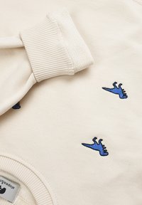 Felpa color crema con polsini a coste e piccole ricamature di dinosauri blu su tutta la superficie. Tessuto morbido, design casual.