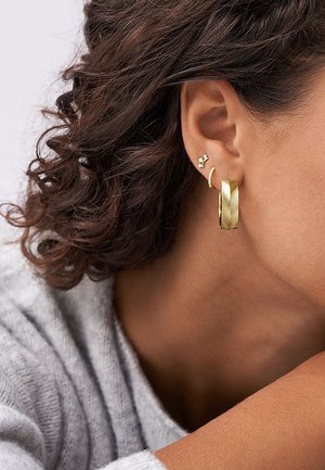 Boucles d'oreilles en or en forme de créoles avec un motif texturé et une petite boucle d'oreille en forme de fleur, portées par un modèle aux cheveux bouclés.