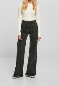 Nainen on pukeutunut valkoiseen ribbitekstiin pitkähihaiseen toppiin, korkeavyötäröisiin, leveälahkeisiin tummanharmaisiin farkkuihin, joissa on etupussit, sekä mustavalkoisiin tennareihin.