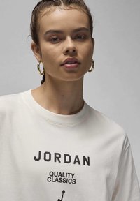 Cream-colored t-shirt featuring a black printed graphic that says "JORDAN QUALITY CLASSICS," with a round neckline and soft texture.