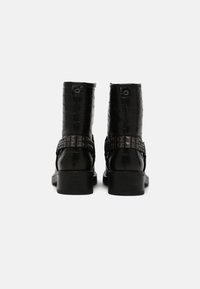 Bottines en cuir noir avec une texture gaufrée croco, featuring une sangle cloutée et un talon carré. Vue positionnée depuis l'arrière.
