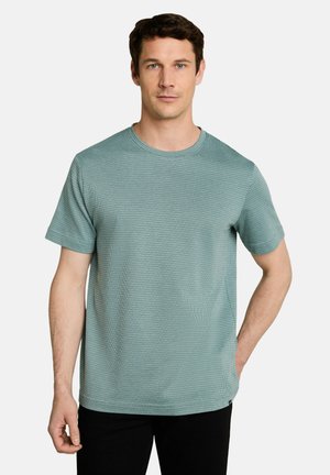 Man met kort donker haar die een gestructureerd lichtgroen shirt met korte mouwen draagt, staand tegen een eenvoudige lichte achtergrond.