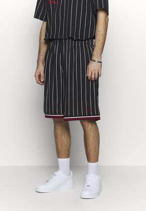 Mand iført sorte ternede shorts og matchende skjorte, hvide sokker og hvide sneakers mod en lys, ensartet baggrund.