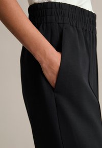 Pantalons noirs avec une taille élastique froncée, dotés de poches latérales. Le tissu semble lisse et légèrement texturé.