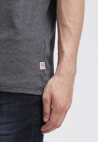 Szara bawełniana koszulka z krótkim rękawem, z małą czerwoną i białą metką marki na dole, w zestawie z ciemnoniebieskimi dżinsami.