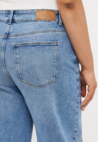 Zilas džinsu bikses ar augstu jostasvietu, aizmugurē esošu kabatas apli un brūnu ādas etiķeti jostasvietā ar iepresētu tekstu.