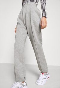 Persona vestida con pantalones de chándal grises de Nike y zapatillas blancas con detalles en azul y rojo, de pie contra un fondo liso.