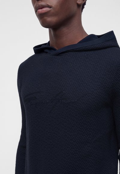 Jeune personne portant un sweat à capuche texturé foncé avec un texte en relief subtil sur la poitrine, montré du menton à la taille sur un fond uni.
