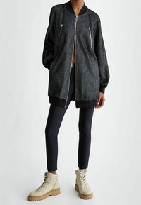 Giacca bomber nera con una finitura testurizzata, tasche con zip e polsini a costine; indossata sopra pantaloni neri aderenti e stivali beige con lacci.