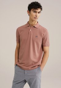 Camisa polo de color rosa claro hecha de algodón, con un cuello de tres botones, mangas cortas y un pequeño logo negro y azul en el pecho.