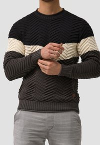 Dunkelgrauer gestrickter Pullover mit einem Zickzackmuster; cremefarbene und schwarze horizontale Streifen; gerippte Bündchen und Saum; kleines Logodetail an der Seite.