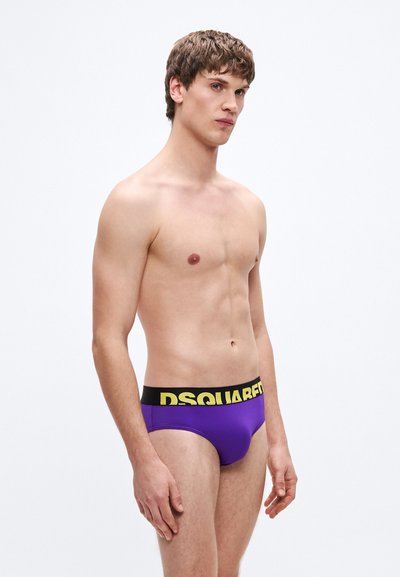 Lila bugyi fekete rugalmas derékpánttal, amelyen sárga "DSQUARED2" logó látható. Simított textúra, testhezálló kivitel, amely kiemeli az alakot.