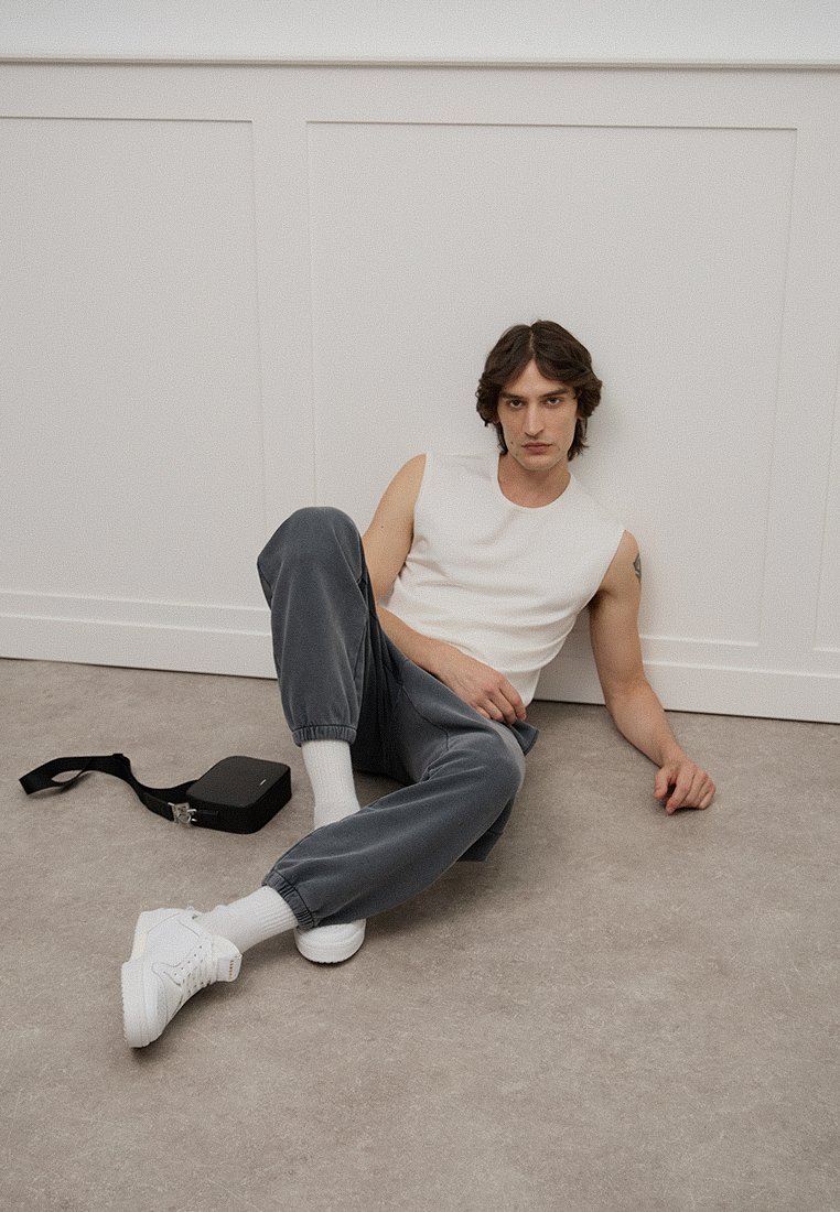 Top sin mangas blanco, joggers gris oscuro, zapatillas blancas y calcetines blancos. Bolsa negra en el suelo. Fondo neutro con elementos de diseño simples.