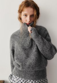 Młoda kobieta z rudymi włosami, ubrana w teksturowany szary sweter na zamek i kraciastą spódnicę, przybliżająca zamek błyskawiczny do twarzy.