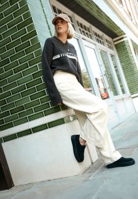 Sorte ruskindssko, cremefarvede bukser med vide ben og en mørkegrå sweater med teksten "SPORTS" i hvid. Miljøet har en væg beklædt med grønne fliser.